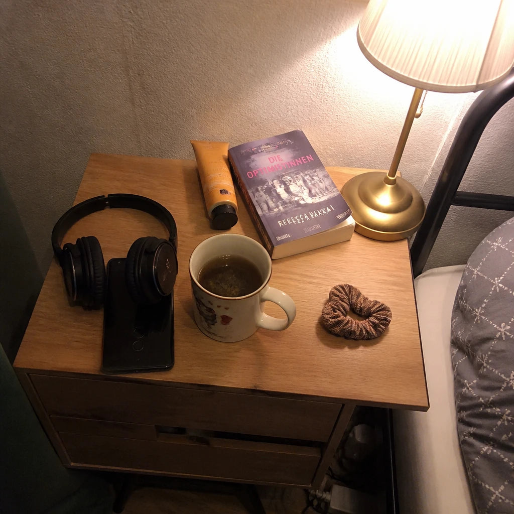 Schnappschuss vom Nachttisch bei warmem Lampenlicht: Kopfhörer auf dem umgedrehten Handy, daneben Tee, Haargummi und ein angelesenes Taschenbuch