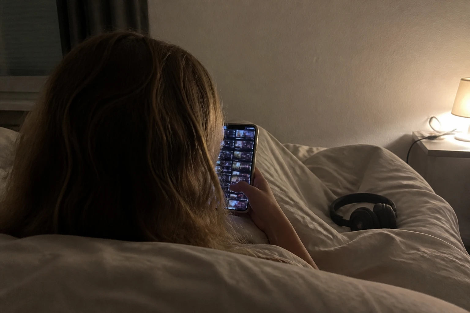 Nachtaufnahme über die Schulter einer Frau im Bett: auf dem Handy eine endlose Liste fast identischer Story-Kacheln, Kopfhörer liegen unbenutzt auf der Decke