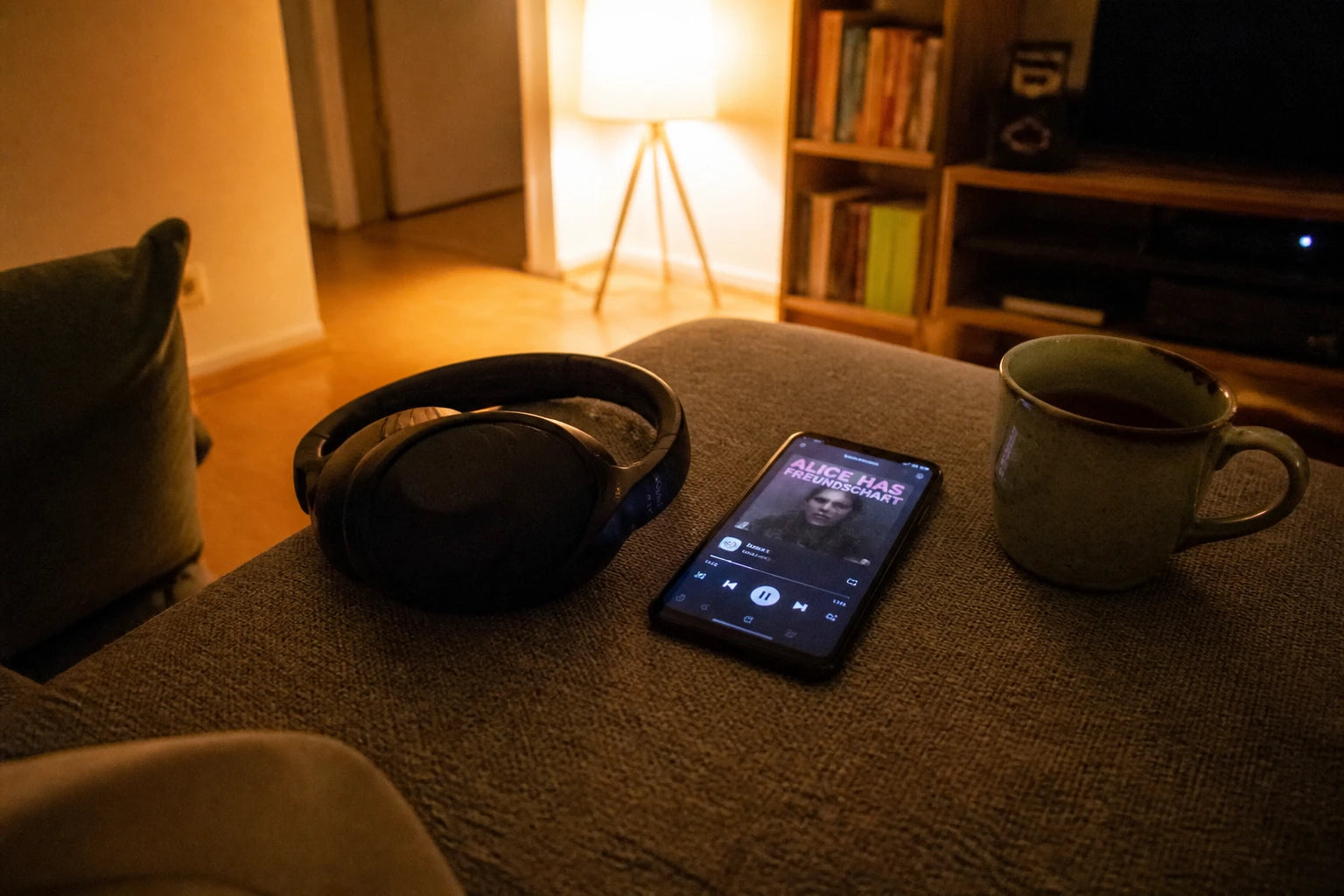 Kopfhörer und Handy mit pausiertem Audio-Player auf einem grauen Sofa, warmes Lampenlicht